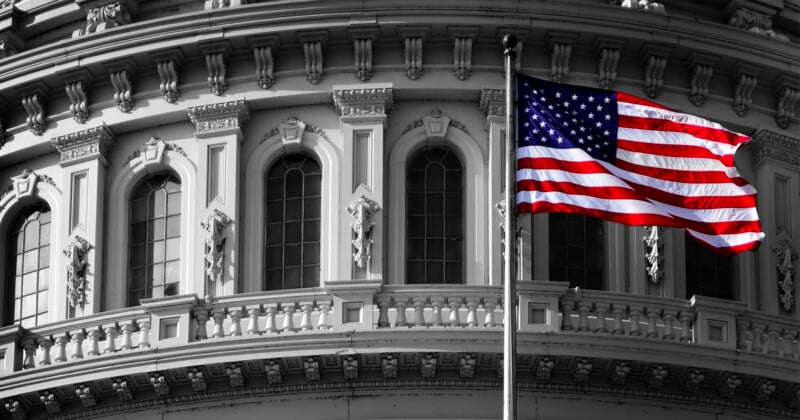 A United States flag waves in front of a portion of a building with classical architectural elements including arched windows and ornate moldings. The image is primarily black and white with the flag in color.