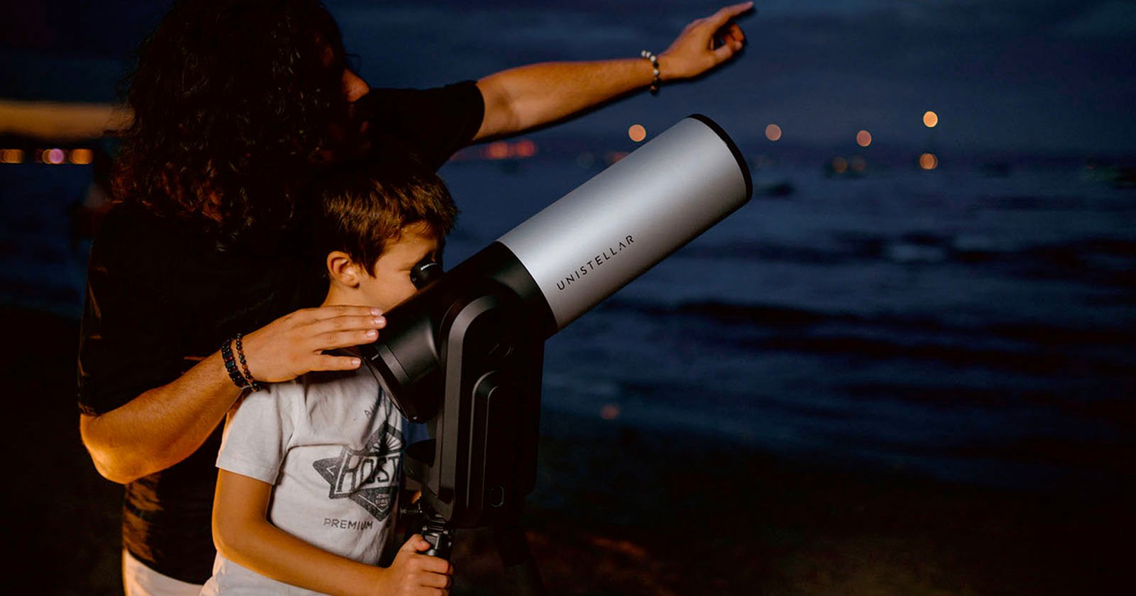 A child looking through a telescope on a beach at night with an adult standing behind and pointing towards the sky. The sea is visible in the background with lights dotting the horizon.