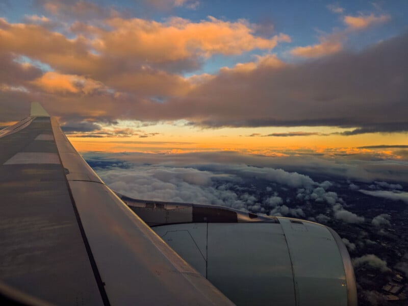 A scenic view from an airplane window, showing the aircraft wing and engine, with a vibrant sky filled with golden and pinkish clouds during sunset. The landscape below features a mix of cloud cover and glimpses of land.