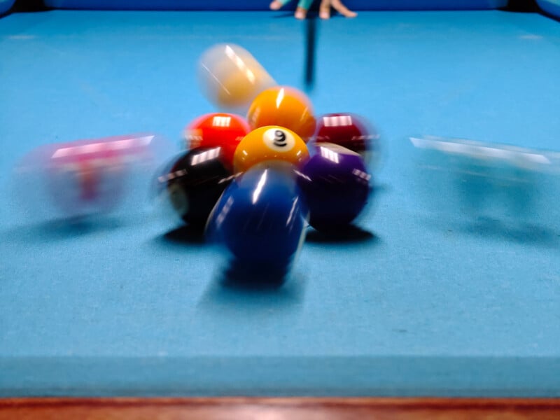 A close-up of a pool table shows a break shot scattering the pool balls in various directions. The cue stick is visible in the background, and the blue felt surface contrasts with the colorful balls in motion.