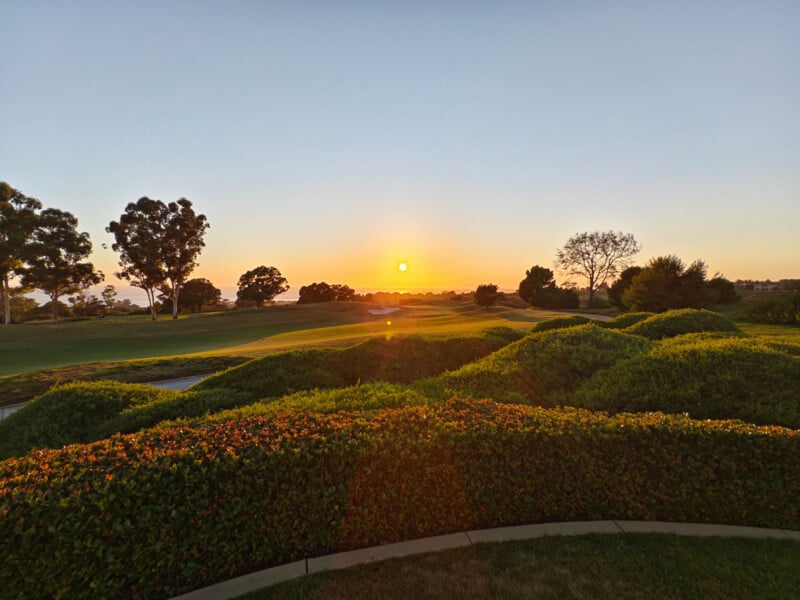 A scenic view of a golf course at dawn, featuring lush, manicured shrubs and expansive green fairways. The sun is rising in the clear sky, casting a warm golden light over the landscape. Tall trees are silhouetted against the horizon.