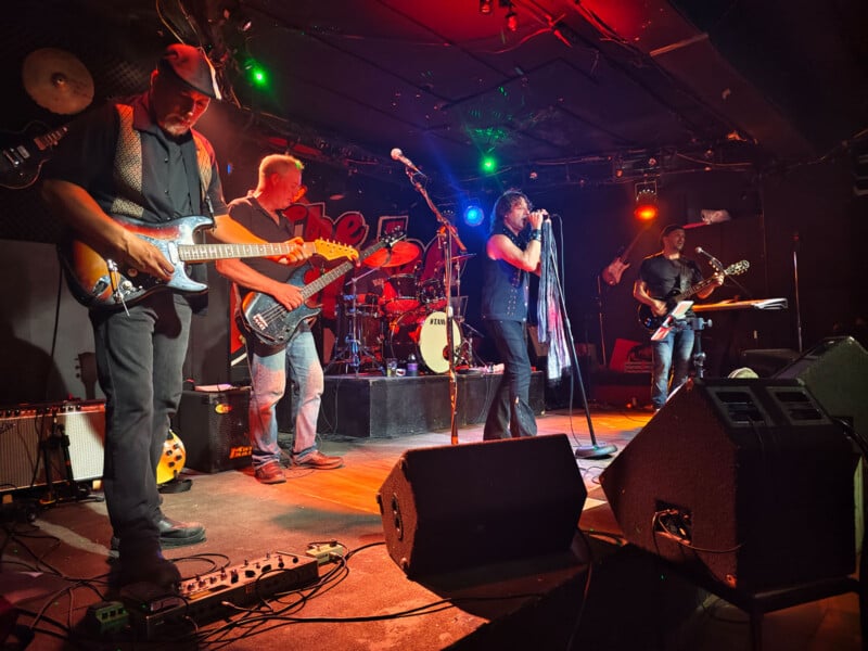 A band performs on stage at a dimly-lit venue with colorful spotlights. The group includes a lead singer, two guitarists, a bassist, and a drummer. The musicians are deeply engaged with their instruments, creating an energetic atmosphere. Various equipment is visible around them.