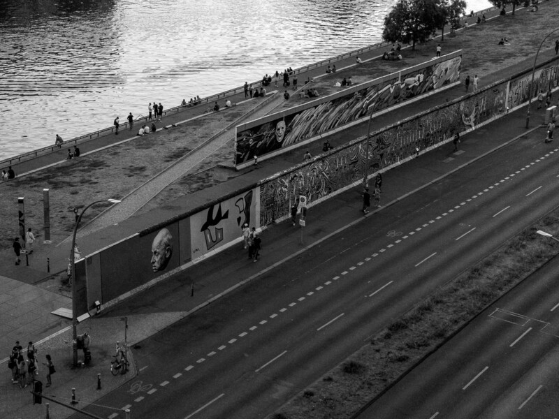 A black and white image of a riverbank with people relaxing on the grassy area. Parallel to the river, there's a long mural on a wall with various artworks. A road runs alongside the wall, with scattered pedestrians and cyclists. Trees dot the far side of the riverbank.