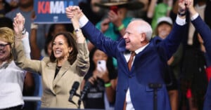 A woman in a tan suit and a man in a blue suit hold hands and raise them high in celebration on a stage, with an enthusiastic crowd in the background. One person holds a sign partially displaying the name "KAMA.