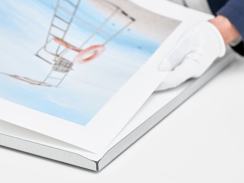 A gloved hand carefully handling a large print of a photograph showing a lifeguard chair and a lifebuoy on a beach. The print is partially rolled or unbent over a white surface. The person appears to be inspecting or preparing the print for framing or packaging.