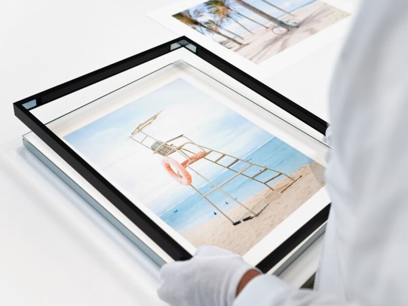 A person wearing white gloves is placing a photograph of a lifeguard stand at a beach into a black picture frame. Another framed photograph of a beach scene with palm trees and a bicycle is visible in the background on the white surface.