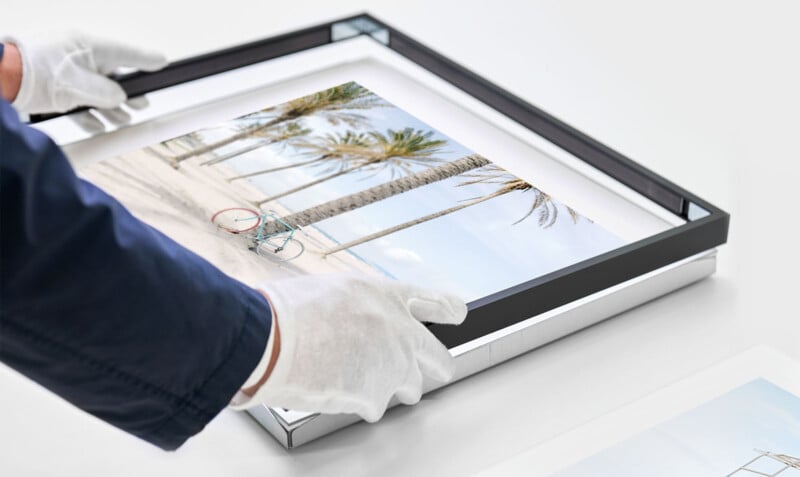 A person wearing white gloves and a dark blue jacket is carefully handling a framed photograph of a beach scene with palm trees and a bicycle. The person is positioning the frame on a white surface.