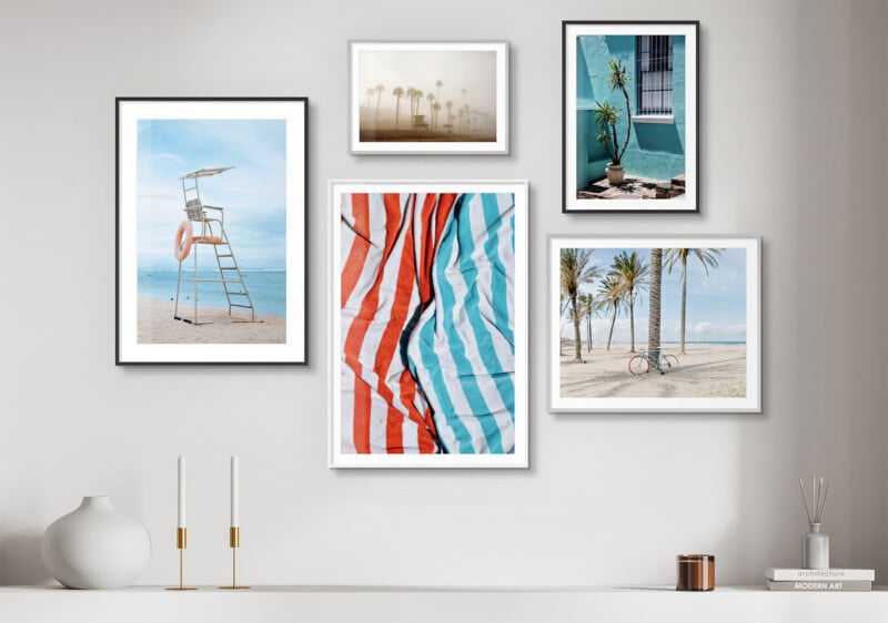 A gallery wall features six framed beach-themed photographs. Images include a lifeguard chair, palm trees in fog, a colorful beach towel, and a bike near palm trees. The frames vary in size and orientation, all displayed on a light grey wall above a minimal shelf.