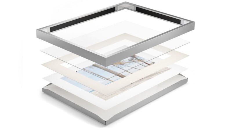 An exploded view of a framed picture showcasing its layers. The frame is silver and rectangular. The layers include glass, a mat with a central opening, a photograph of palm trees and a beach, and a solid backing. Each layer is separated and floating above one another.