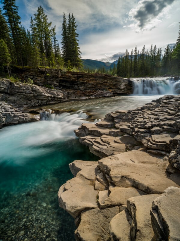 A scenic view of a waterfall cascading over layered rock formations into a clear, turquoise pool, surrounded by dense evergreen trees under a partly cloudy sky, with distant mountains visible in the background.