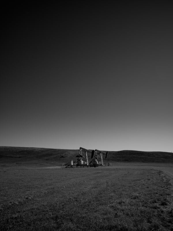 Black and white photograph of an isolated landscape showing two oil pump jacks in the middle of a vast, grassy field under a clear sky. Simple terrain with no trees or additional structures visible, accentuating the solitude and industrial presence.