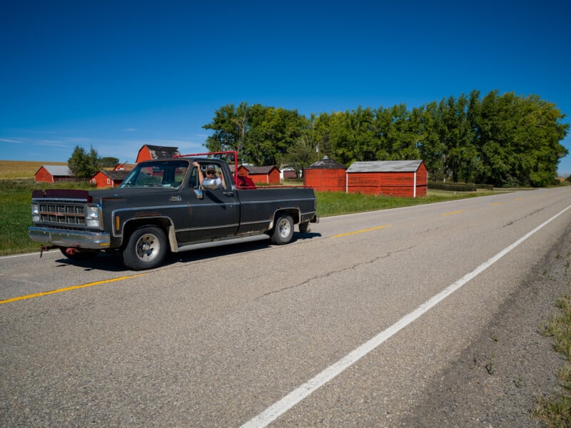 A classic black pickup truck with a red interior is parked on the side of a rural road with scattered red barns and farm buildings in the background. The scene is set on a sunny day with a clear blue sky and lush green trees.