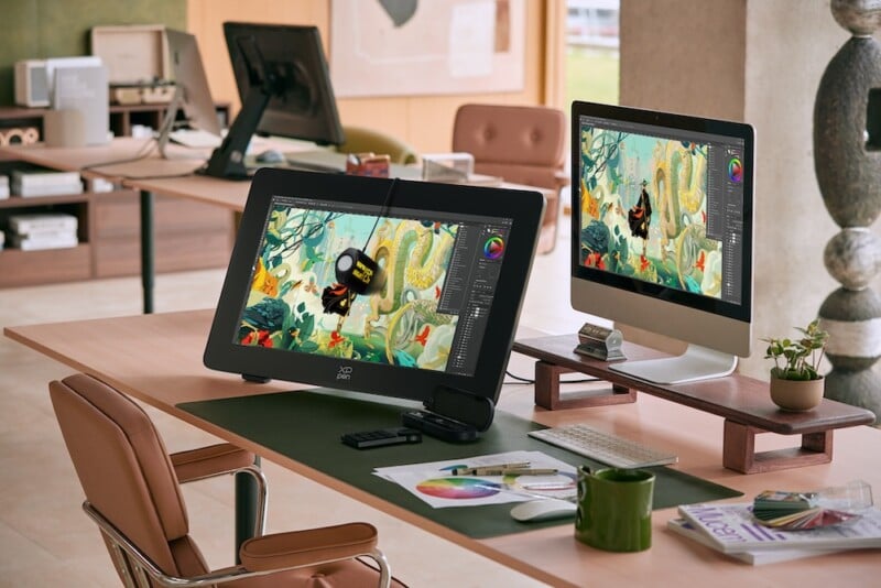 A modern office desk setup featuring a drawing tablet and a monitor, both displaying colorful digital artwork. The desk also has a swivel chair, a green desk mat, a mug, drawing tools, and neatly arranged books and papers. An open-plan office is visible in the background.