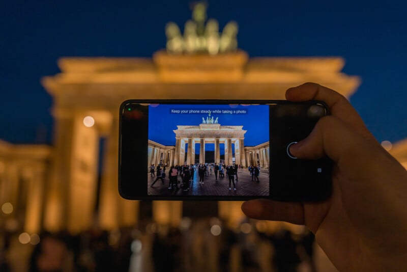 A hand holds a smartphone up against the nighttime backdrop of the illuminated Brandenburg Gate in Berlin, capturing the iconic structure on the phone's screen. The scene features numerous people gathered around the gate, creating a lively atmosphere.