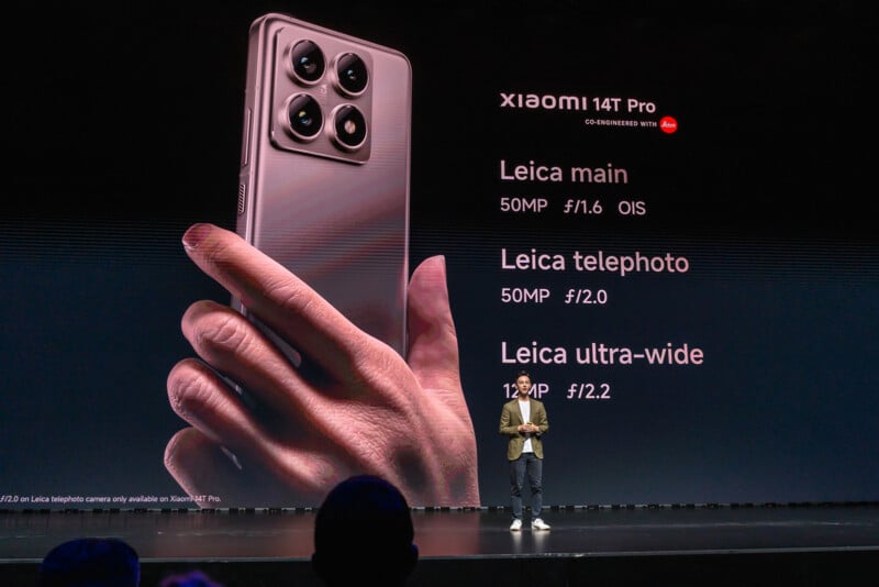 A person stands on a stage presenting the Xiaomi 14T Pro smartphone, which is displayed on a large screen behind them. The screen highlights the phone's camera specs: 50MP main camera with f/1.6 OIS, 50MP telephoto with f/1.9, and 12MP ultra-wide with f/2.2.