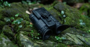 A pair of black binoculars rests on moss-covered rocks and surrounded by small plants. The binoculars have a sleek design with various buttons and a central hinge, situated in a lush, green outdoor environment.