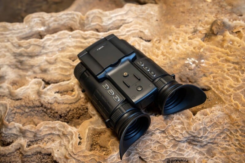 A black dual-lens night vision binocular device rests on a rough, textured beige surface, likely rock or coral. The device features various buttons and dials, with eyepieces on one end and a rectangular back.