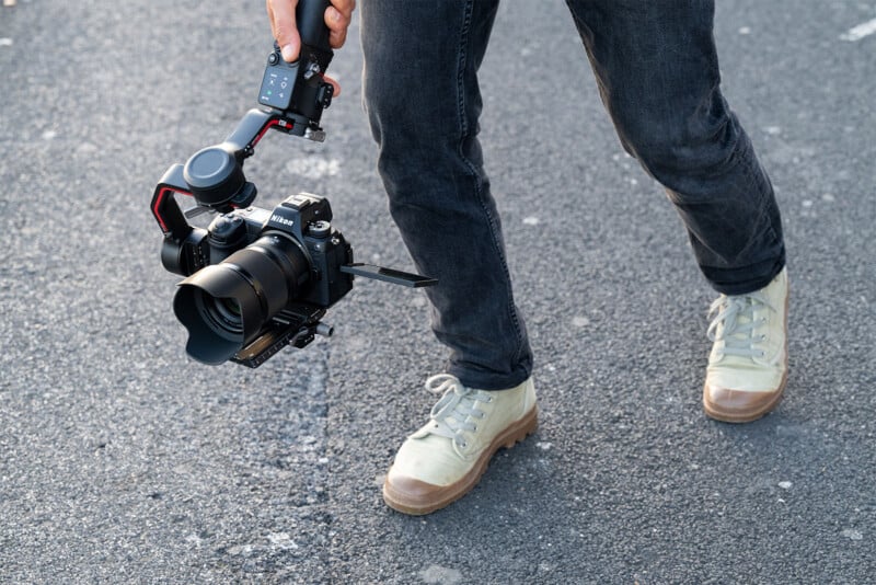 A person holding a professional DSLR camera mounted on a gimbal stabilizer. The person is wearing light-colored sneakers and dark jeans, and is walking on a textured asphalt surface. The camera and gimbal are positioned low to the ground.