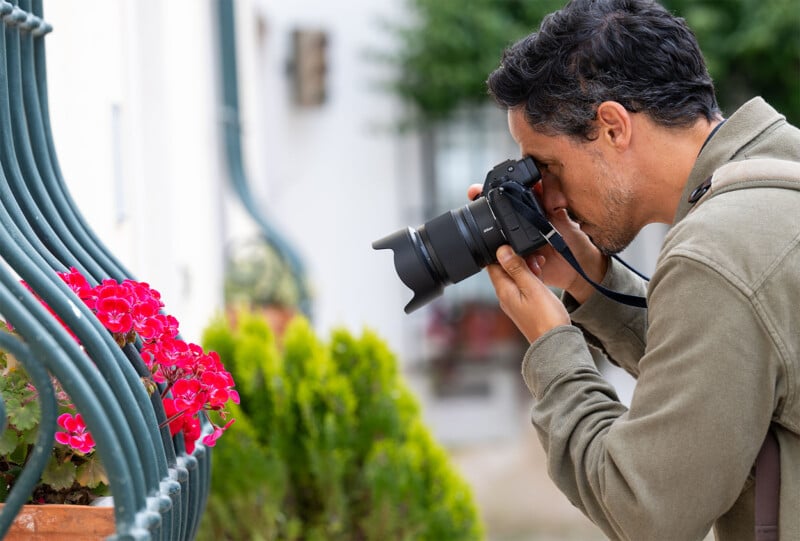 A man with short dark hair and a beard is using a camera to photograph bright red flowers in a garden. He is holding the camera close to his face and wearing a khaki jacket, with greenery and blurred background visible.