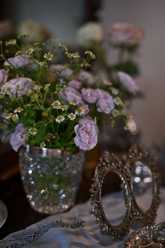 A crystal vase filled with light purple roses and small white flowers is placed on a lace-covered table. Beside the vase, two ornate, vintage-style photo frames are visible, adding to the elegant and nostalgic atmosphere of the setting.