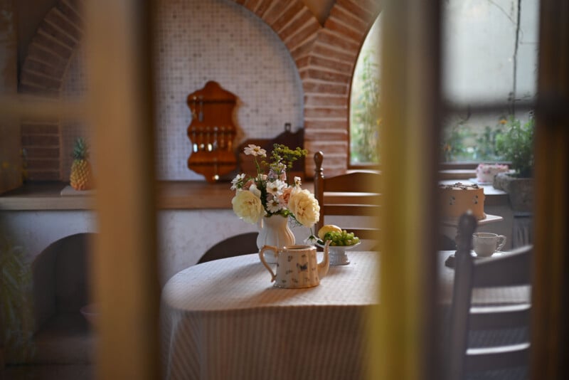 A cozy kitchen features a table set with a floral centerpiece, a ceramic jug, and a bowl of green apples. The background includes a tiled wall, a wooden utensil holder, and arch windows, creating a warm and inviting atmosphere.