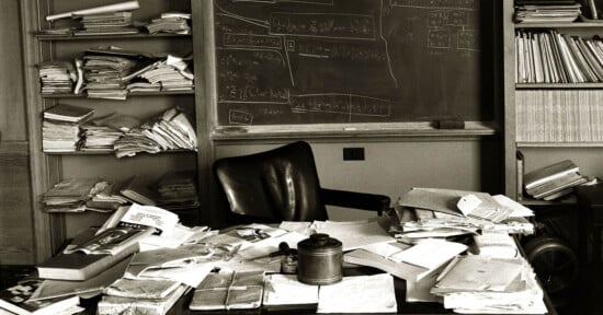 A cluttered office desk filled with scattered papers, books, and folders sits in front of a chalkboard covered with mathematical equations. Shelves in the background are stacked high with more disorganized papers and files. An empty chair is situated behind the desk.