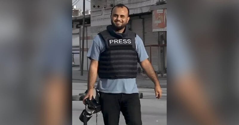 A man wearing a blue shirt and black bulletproof vest labeled "PRESS" stands outdoors, smiling, and holding a camera. The background features closed shop fronts and a gray street. He appears to be in a professional setting, possibly reporting or documenting an event.