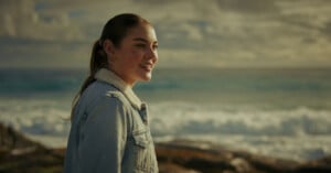 A woman with her hair tied back, wearing a light denim jacket with a fuzzy collar, smiles while standing by the seaside. The background features a scenic ocean view with waves crashing and a cloudy sky.