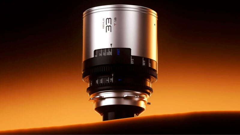 A silver camera lens with black detailing and various markings, including "33" and "T1.3," is centered in the image. The lens appears to be resting on a surface with a warm, orange backlight creating a soft glow and gradients in the background.