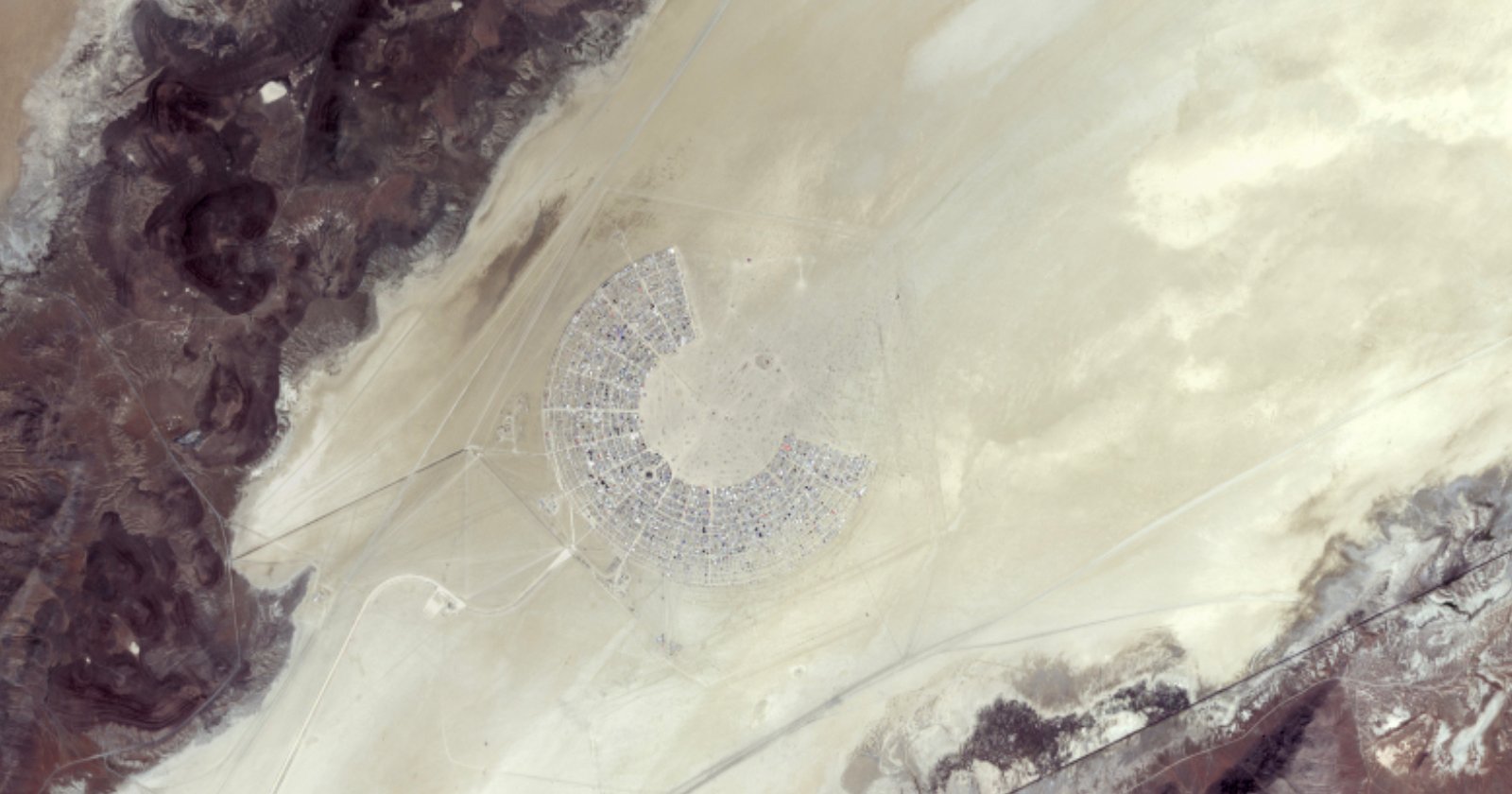 A satellite image of a large, organized desert settlement arranged in a semi-circular pattern, surrounded by vast stretches of barren land. The settlement has a distinct grid layout with numerous roads and pathways. Darker terrain is visible on the fringes.