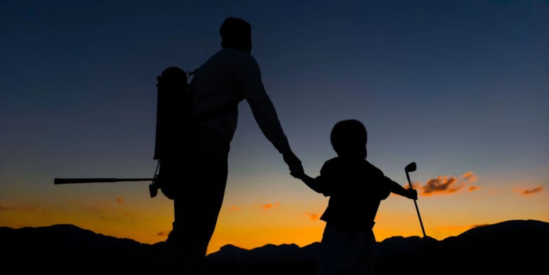 A silhouetted adult and child holding hands while standing on a golf course during sunset. The adult carries a golf bag, and the child holds a golf club. The sky is a gradient of deep blue to warm orange, with mountains in the background.