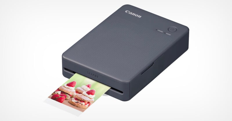 A compact Canon photo printer with polygonal pattern on top is printing an image of dessert with strawberries and cream. The small printer is designed for printing high-quality photos and features a minimalistic design in a dark grey color.