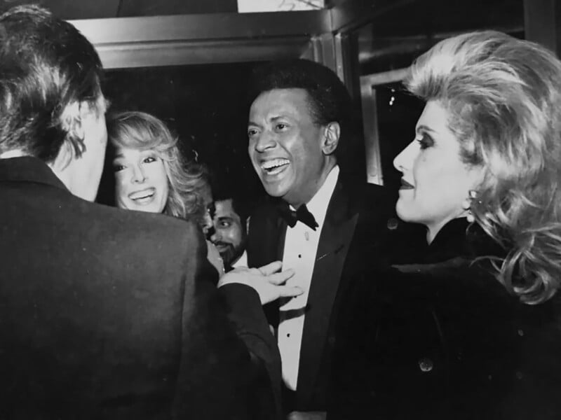 A black-and-white image showing a group of people dressed in formal attire engaged in conversation. One man in a tuxedo is smiling brightly, while two women with voluminous hairstyles also appear to be enjoying the interaction.