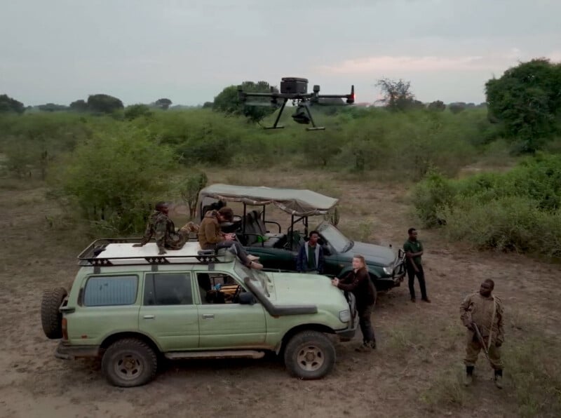 A group of people stands around two off-road vehicles in a grassy, open area. One person is on top of a vehicle looking up at a large drone flying above them. The sky is overcast, and the surrounding area is filled with green bushes and trees.