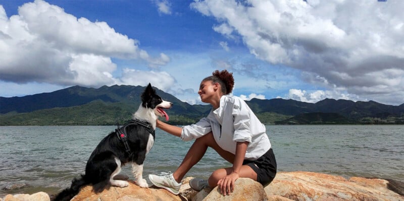 A woman sits on rocks by a lake, smiling and petting a black and white dog. The woman wears a white shirt and black shorts, and the dog is on a leash. Mountains, trees, and a partly cloudy sky are visible in the background.