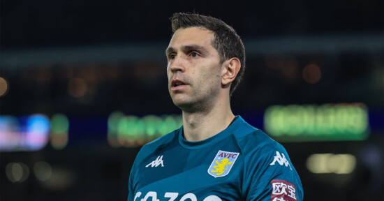 A soccer player in a teal uniform with logos including "Cazoo" on the front and the Aston Villa crest, is standing on the field. The player appears focused, with a blurred stadium backdrop behind him.