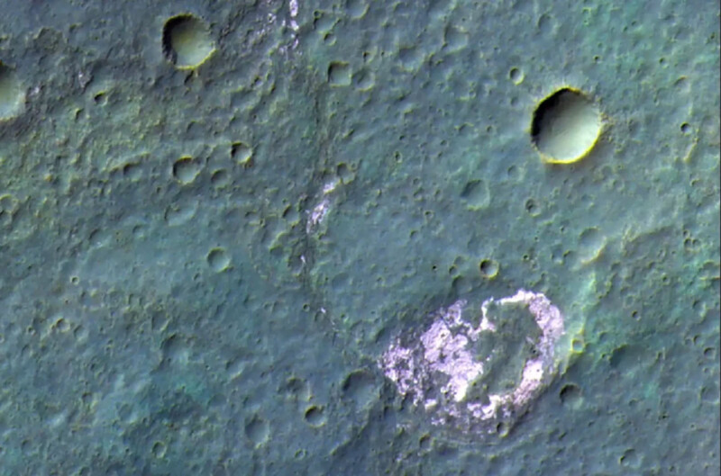 A high-resolution image of the Moon's surface showing various craters of different sizes. The terrain appears rugged, with a large, prominent, white-colored crater at the lower right side, contrasting with the darker, greenish surrounding areas.