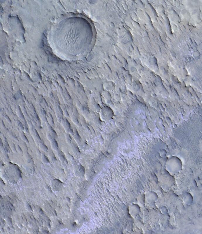 Aerial view of a Martian landscape featuring several impact craters of varying sizes. The surface has a rugged, ridged texture with streaks of blue-tinted material. The largest crater is near the top left corner, showing a smoother basin in the center.