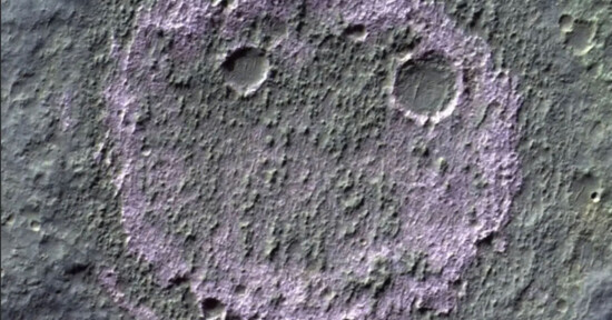 Aerial view of a cratered section of the Martian surface, where three distinct craters form a pattern resembling a smiling face. The "eyes" are two small circular craters, and the "mouth" is a larger, curved crater below them. The surface shows textured terrain.