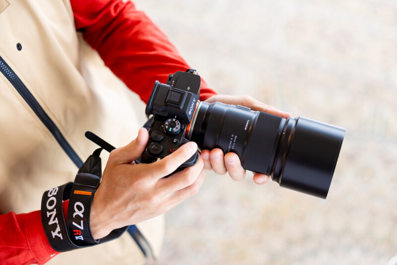 Person in a beige and red jacket holding a Sony Alpha 7R IV mirrorless camera with a large lens, adjusting settings. The camera strap around their wrist is marked with "α7R IV" and "Sony". The background is blurred.