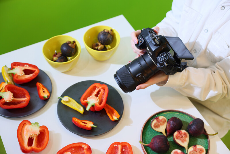 A person in a white shirt is taking a picture of cut bell peppers and figs on plates with a camera. The table is also adorned with two yellow bowls filled with figs against a vibrant green background.