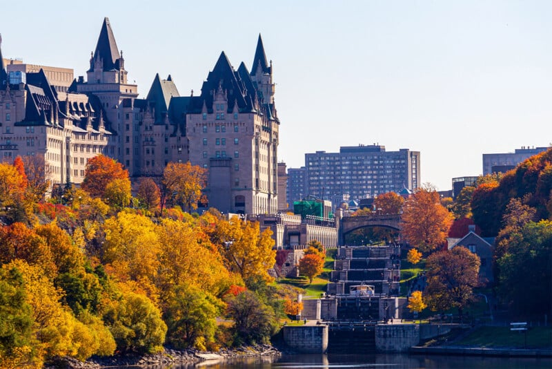 A scenic view of a historic castle-like building surrounded by vibrant autumn foliage. Below, a series of locks leads down to a river, with the cityscape featuring a mix of older and modern buildings in the background under a clear blue sky.