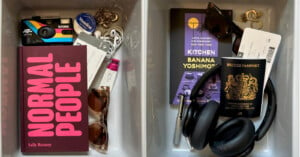 Two trays at an airport security checkpoint. The left tray contains a book titled "Normal People," sunglasses, a keychain, wired earphones, and miscellaneous small items. The right tray holds another book, a pair of sunglasses, a British passport, headphones, and a boarding pass.