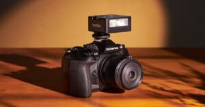 A black Olympus camera is placed on a wooden surface with a warm yellow background. The camera is equipped with a Godox flash mounted on top. Shadows and light create a dramatic effect around the camera.