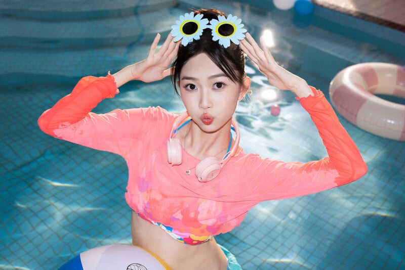 A young woman stands in a pool with her arms raised beside her head. She has a playful expression, pursing her lips, and wears a bright floral top, pink headphones around her neck, and a pair of decorative sunglasses with flower frames. A beach ball floats near her.