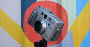 A silver Canon PowerShot G2 digital camera mounted on a tripod in front of a colorful background with red, blue, yellow, and green abstract shapes. The camera's lens and various buttons are visible.