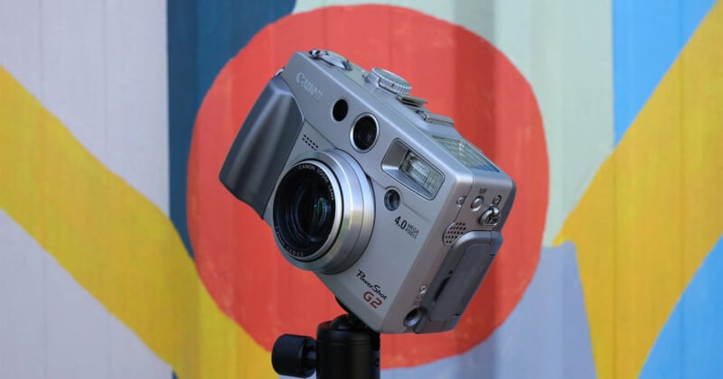 A silver Canon PowerShot G2 digital camera mounted on a tripod in front of a colorful background with red, blue, yellow, and green abstract shapes. The camera's lens and various buttons are visible.