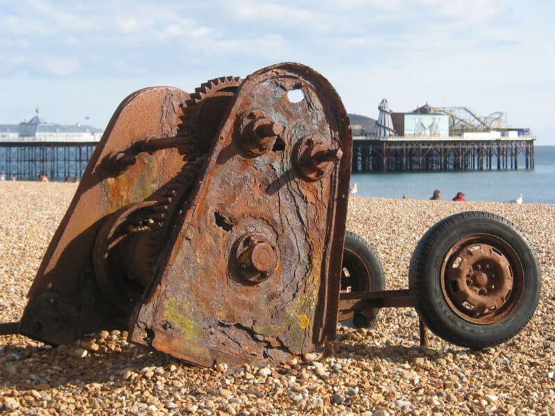 An old, rusted mechanical structure lies on a pebble beach with two wheels attached. In the background, there's a pier stretching into the water with a few people and seagulls scattered on the beach. The sky is partly cloudy.