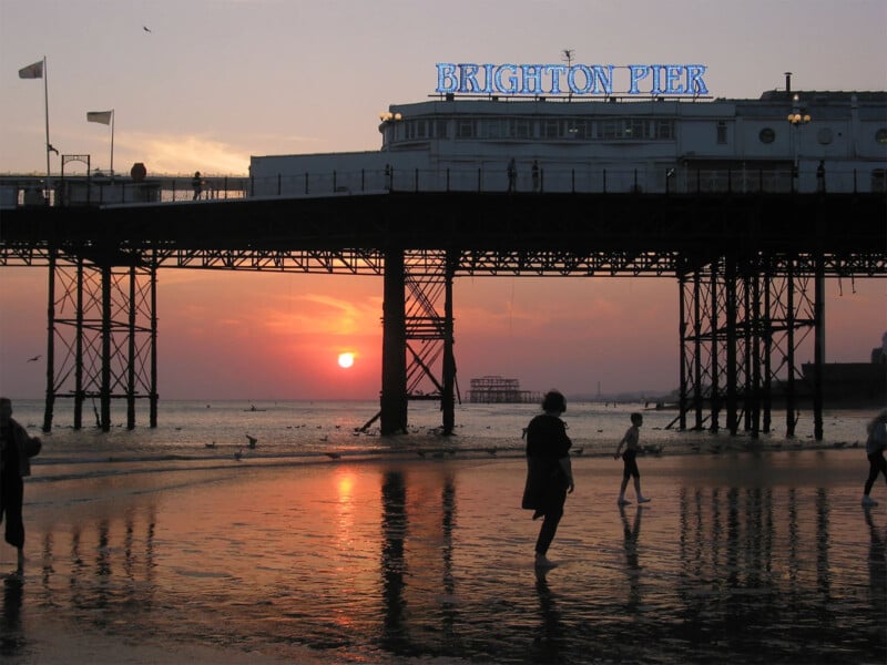 People walking on the beach at sunset with the Brighton Pier structure in the background. The sky is filled with warm hues of orange and pink, and the sea reflects the setting sun. The neon sign of "Brighton Pier" is visible at the top of the pier.