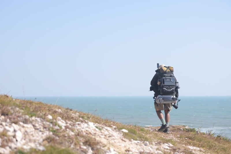 A person is hiking along a grassy and rocky cliffside path with a large backpack on a clear, sunny day. The ocean extends into the horizon under a pale blue sky.